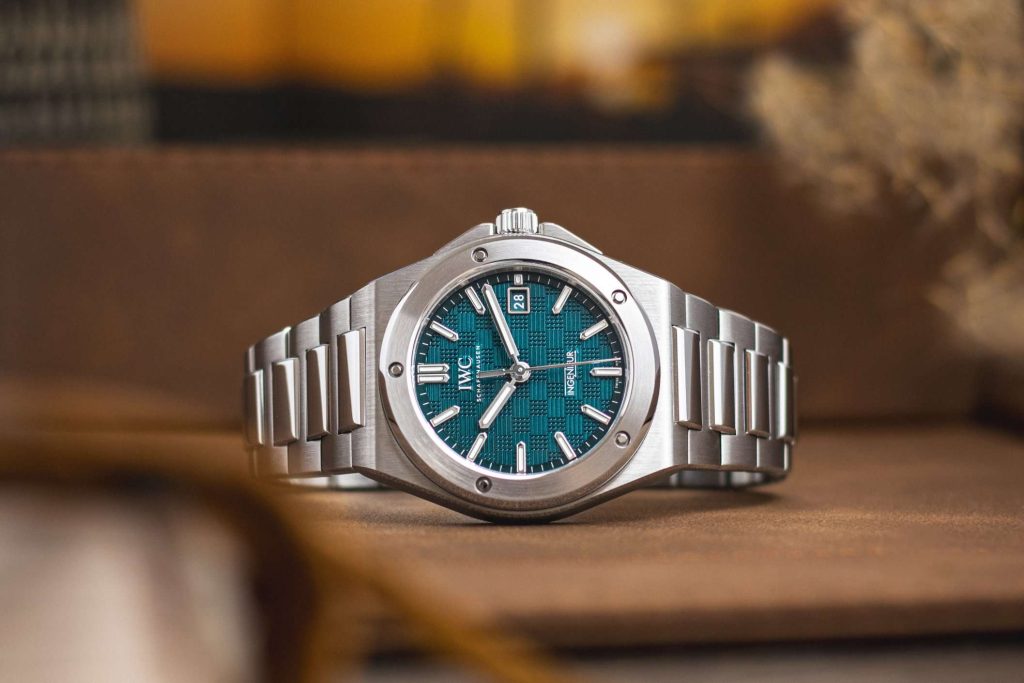 IWC Ingenieur mit Petrol-Zifferblatt liegend auf einem Untergrund aus Holz mit Gegenständen im Vorder- und Hintergrund, die ebenfalls in warmen Farben dargestellt sind