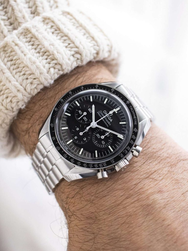 OMEGA Speedmaster Professional Moonwatch am Edelstahlarmband abgebildet an einem Handgelenk mit beigem Ärmel vor weißem Hintergrund