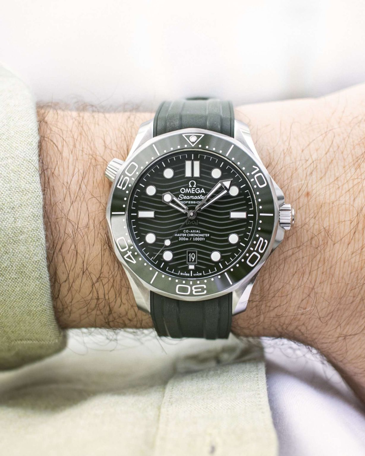 Omega Seamaster Diver 300M mit grünem Zifferblatt, grüner Lünette und grünem Kautschukband abgebildet an einem Handgelenk mit hellgrünem Ärmel vor weißem Hintergrund