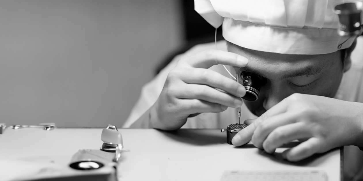 Ein Handwerker von Grand Seiko arbeitet mit einer Lupe vor dem Auge an einem Uhrwerk, das zwischen seinen Händen liegt