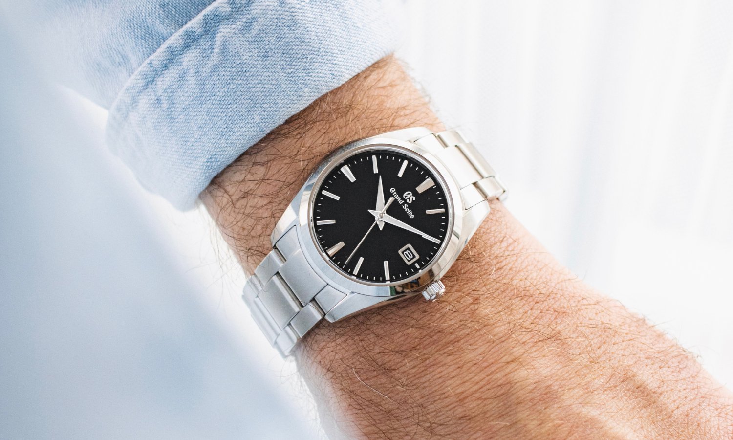 Grand Seiko Heritage Quarz Referenz SBGX261 mit schwarzem Zifferblatt am Edelstahlarmband an einem Handgelenk mit hellblauem Ärmel getragen vor hellem Hintergrund
