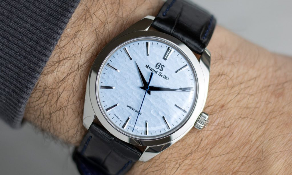 Grand Seiko Omiwatari Referenz SBGY007 mit hellblauem Zifferblatt am schwarzen Lederarmband an einem Handgelenk getragen vor hellgrauem Hintergrund