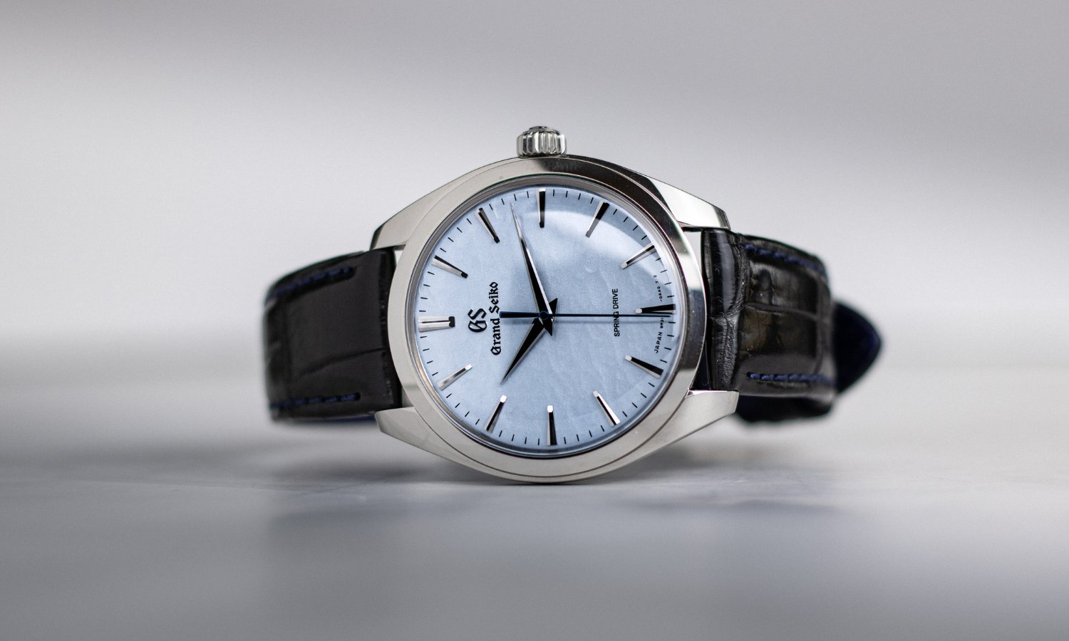 Grand Seiko Omiwatari Referenz SBGY007 mit hellblauem Zifferblatt am schwarzen Lederarmband auf der Seite liegend vor hellgrauem Hintergrund