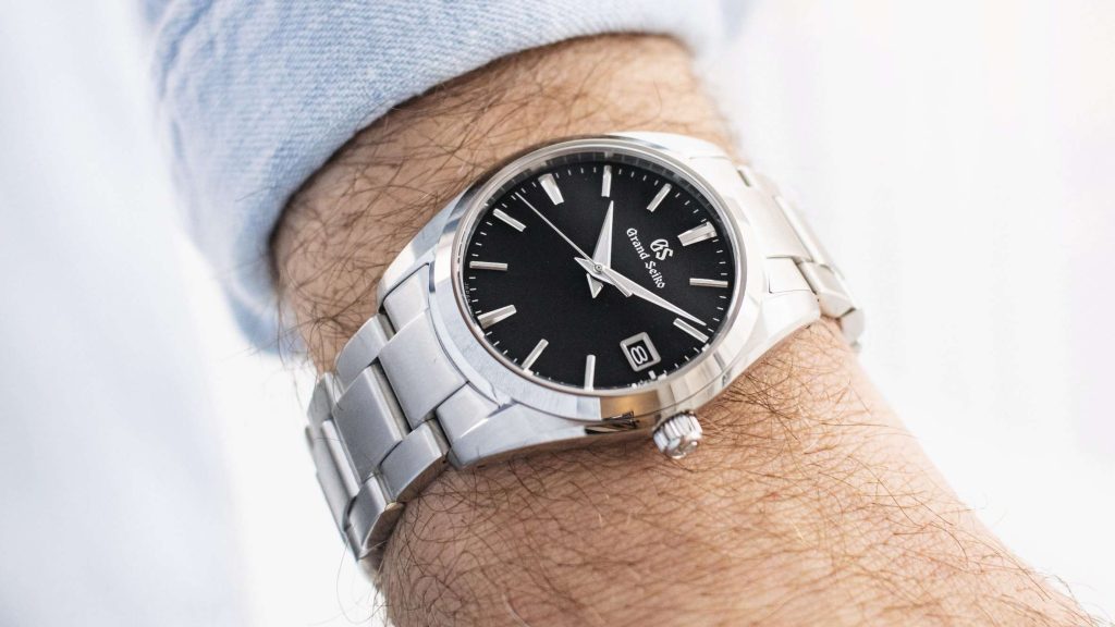 Grand Seiko SBGX261 mit schwarzem Zifferblatt mit Edelstahlarmband abgebildet an einem Handgelenk mit hellblauem Ärmel vor weißem Hintergrund
