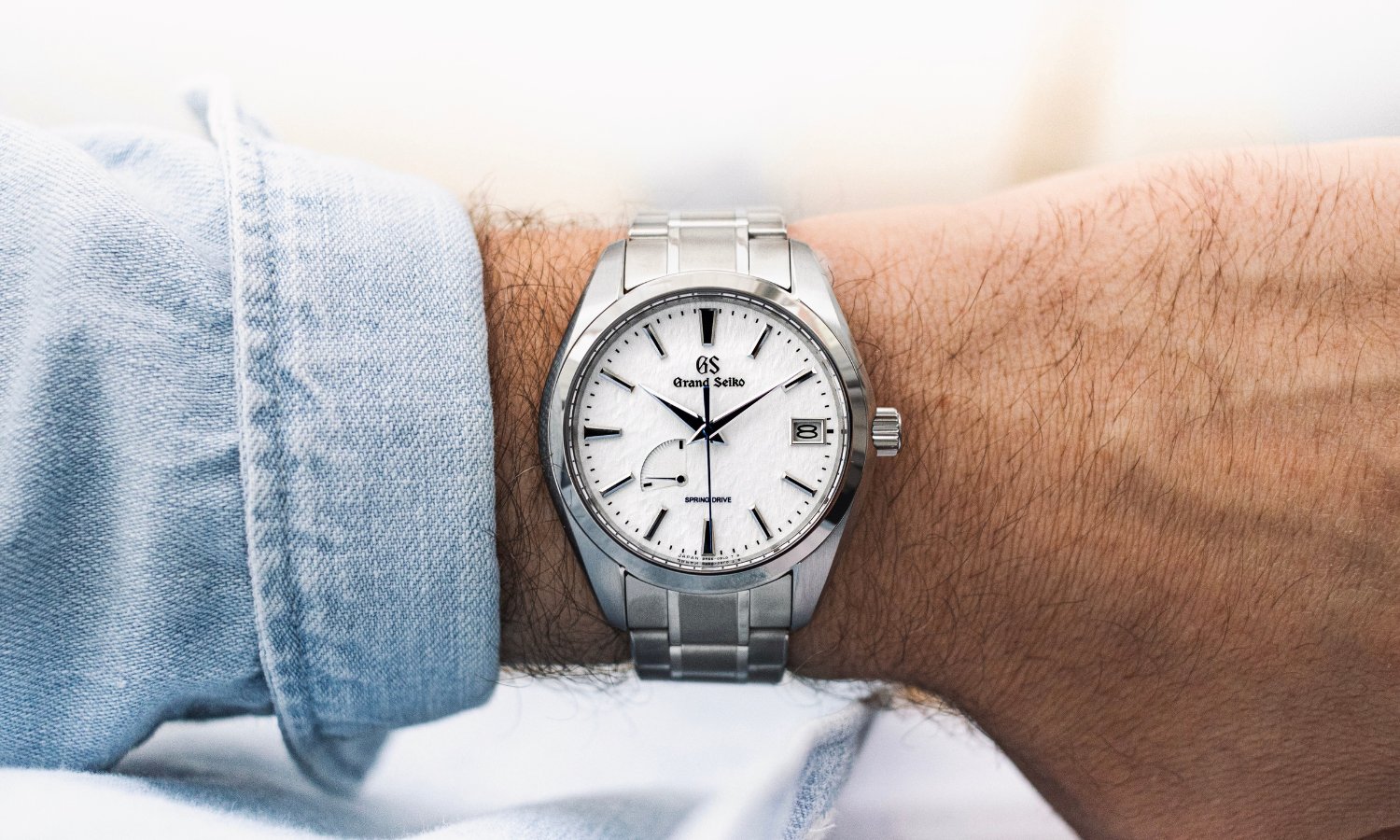 Grand Seiko Snowflake Referenz SBGA211 mit weißem Zifferblatt am Edelstahlarmband an einem Handgelenk mit hellblauem Ärmel getragen vor hellem Hintergrund