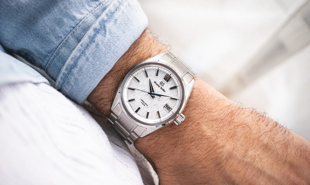 Grand Seiko White Birch Referenz SLGA009 mit weißem Zifferblatt am Edelstahlarmband an einem Handgelenk mit hellblauem Ärmel getragen vor hellem Hintergrund