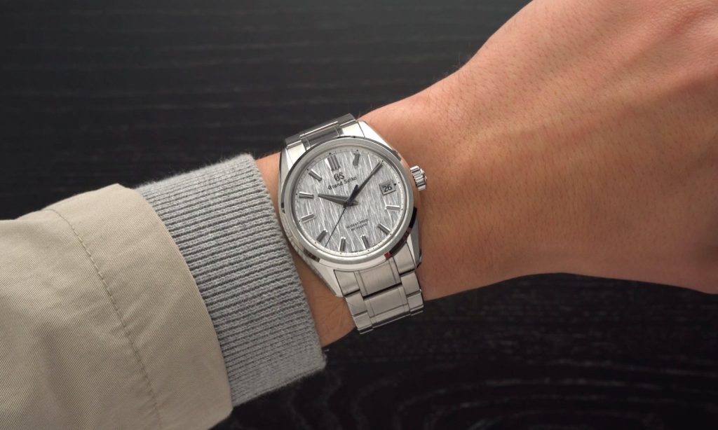 Grand Seiko White Birch Referenz SLGH005 mit weißem Zifferblatt am Edelstahlarmband an einem Handgelenk mit beige grauem Ärmel getragen vor schwarzem Hintergrund