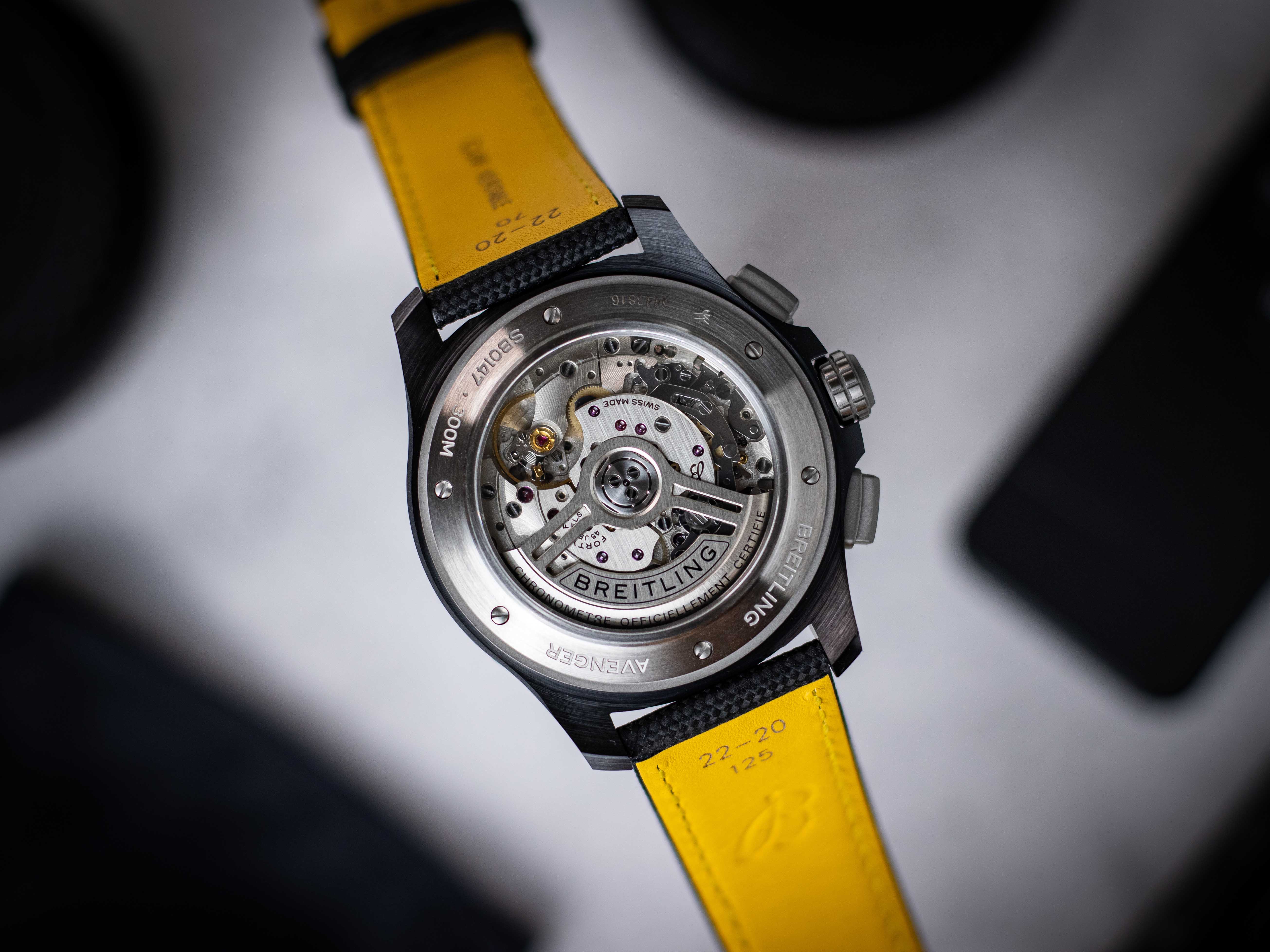 Aufnahme des offenen Gehäusebodens der Breitling Avenger Night Mission Chronograph vor weißem Hintergrund mit schwarzen Gegenständen und einem auf der Rückseite gelben Armband