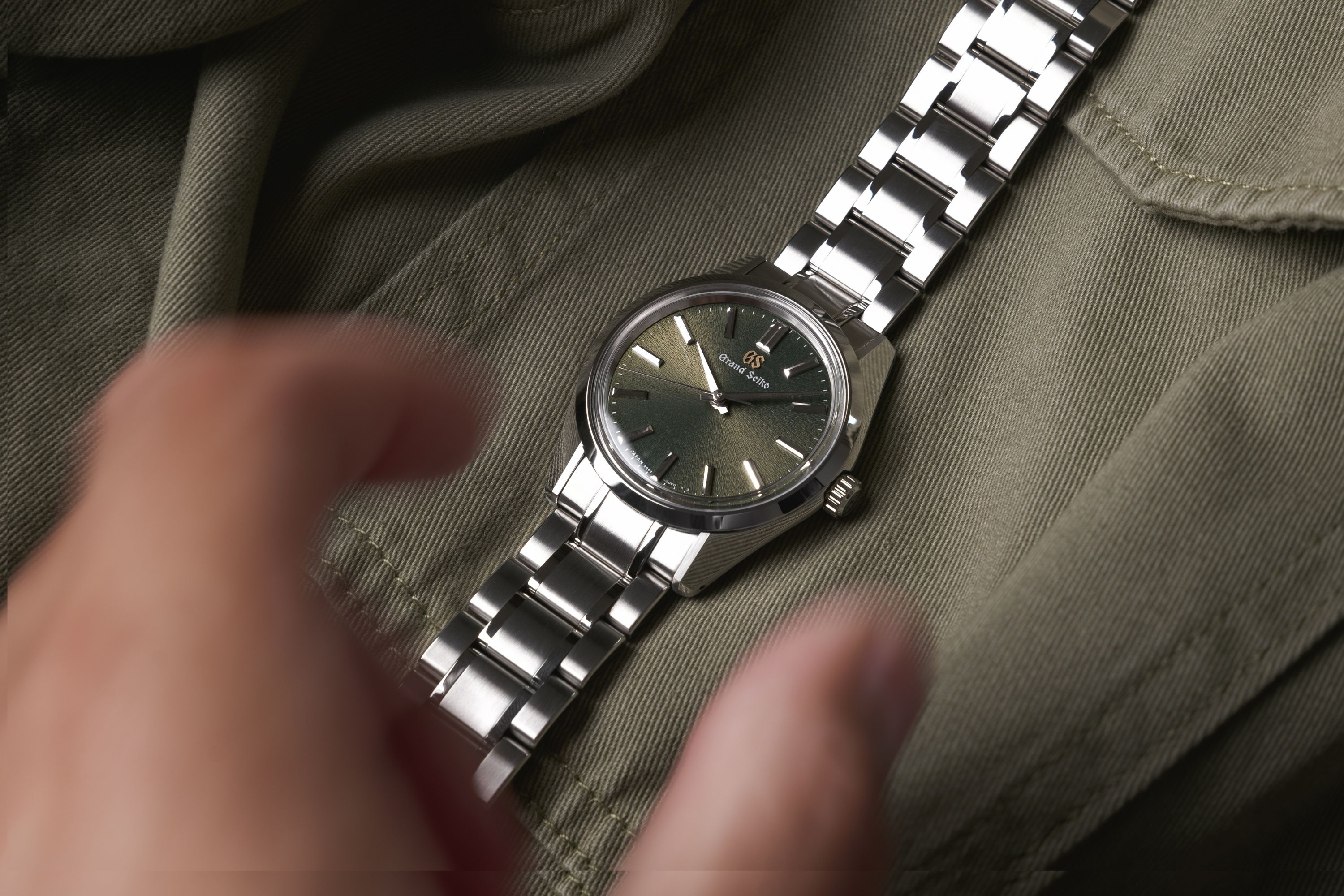Grand Seiko SBGW303 mit grünem Zifferblatt vor grünem Hintergrund zusammen mit einer Hand, die nach der Uhr greift
