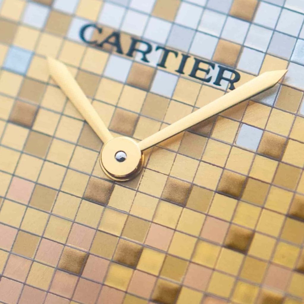 Nahaufnahme der goldenen Schwertzeiger einer Cartier Tank mit kleinen goldblauen Quadraten im Hintergrund