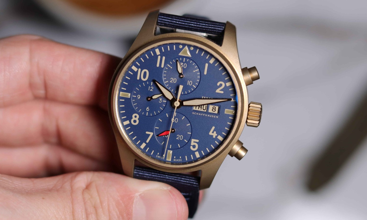 IWC Pilot´s Watch Chronograph in Bronze mit blauem Zifferblatt und blauem Armband gehalten von einer Hand vor weißem Hintergrund mit Gegenständen