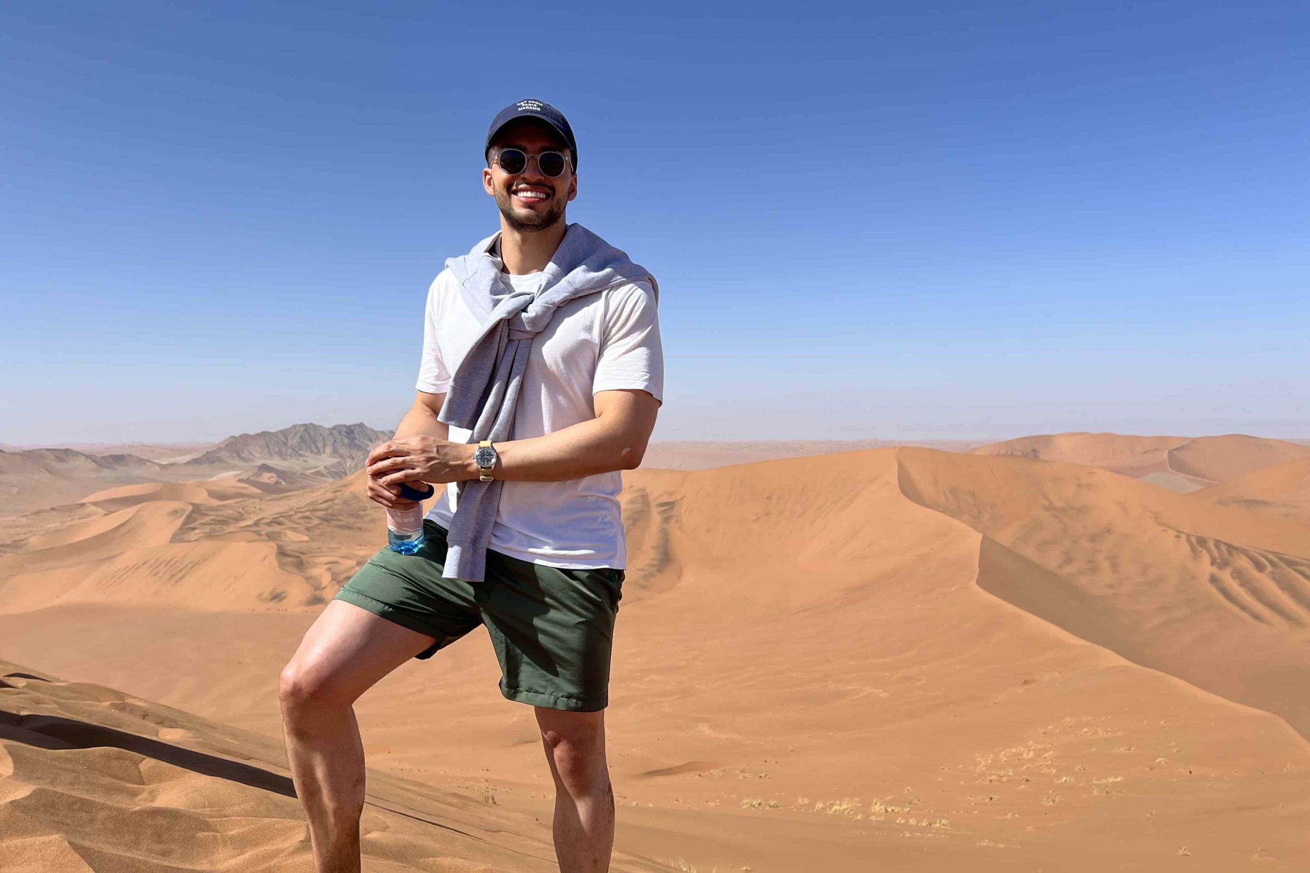 Esteban Montoya mit weißem T-Shirt und dunkler Shorts in der Wüste. Er trägt eine Sonnenbrille, eine Kappe, seine Omega Railmaster und hat eine Wasserflasche in der Hand