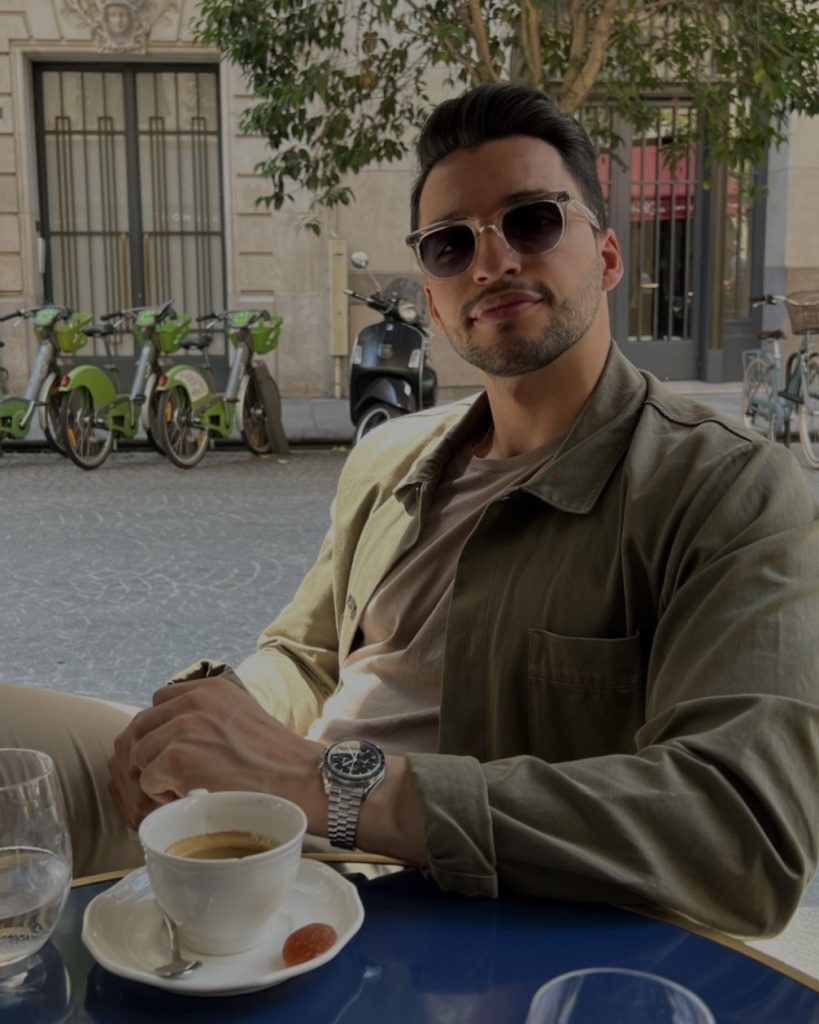 Esteban Montoya sitzt draußen vor einem Café und trinkt einen Espresso. Er trägt eine Sonnenbrille, eine grüne Jacke und seine Omega Speedmaster Professional am Arm