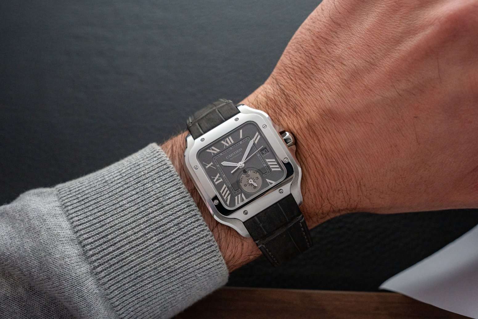 Cartier Santos Dual Time mit grauem Zifferblatt am schwarzen Lederarmband abgebildet an einem Handgelenk mit grauem Ärmel vor dunklem Hintergrund