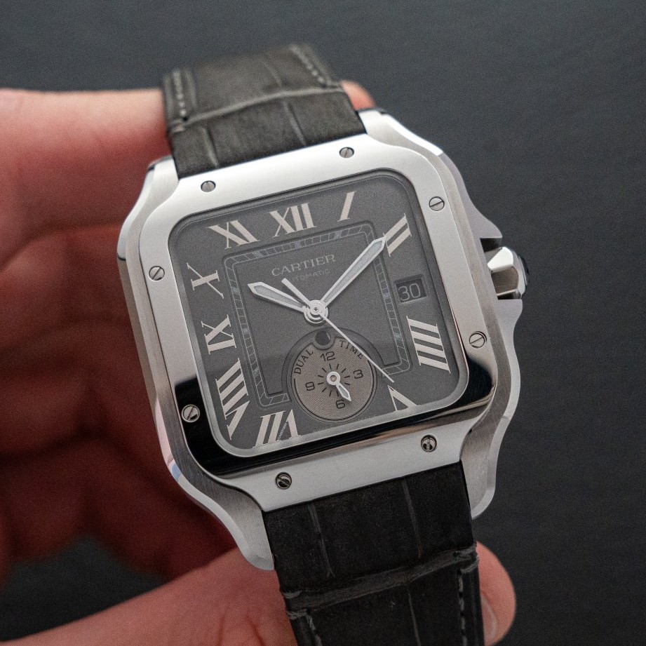 Cartier Santos Dual Time mit grauem Zifferblatt am schwarzen Lederarmband gehalten von einer Hand vor schwarzem Hintergrund