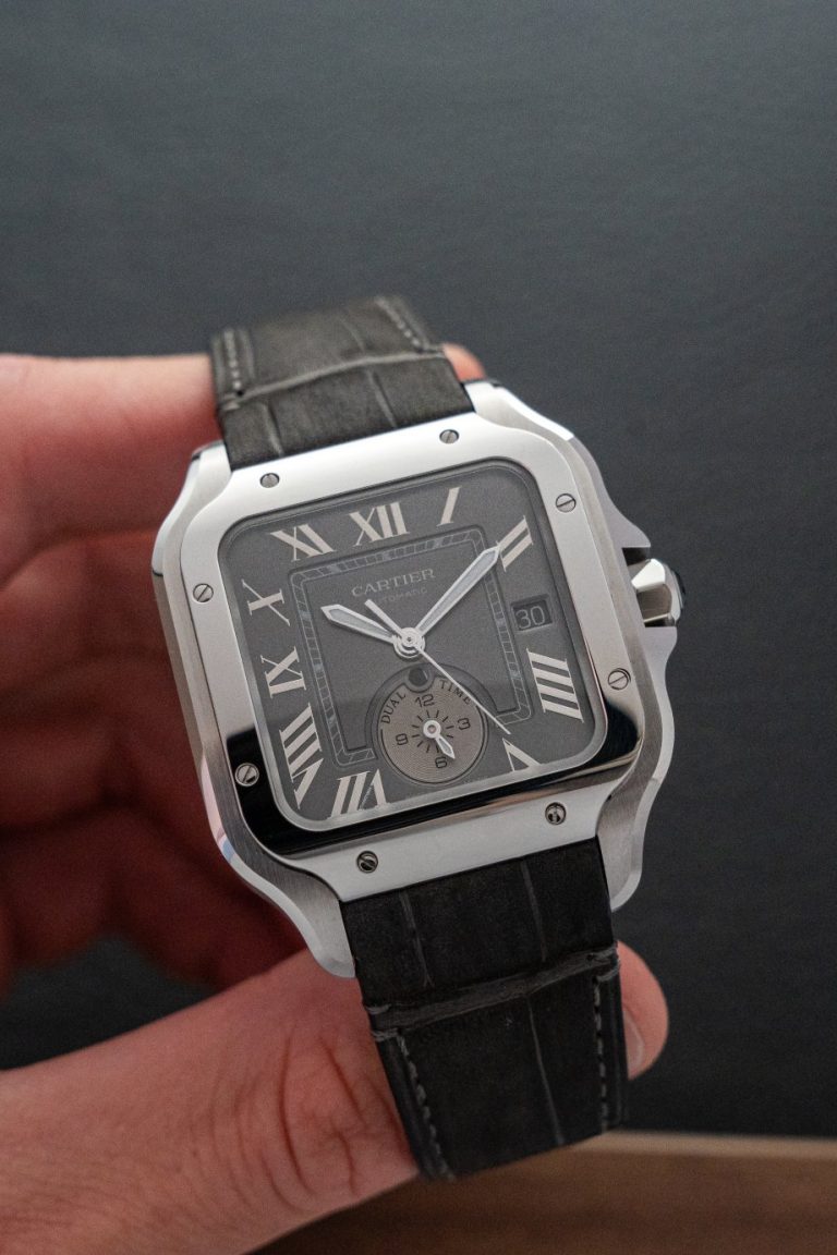 Cartier Santos Dual Time mit grauem Zifferblatt und schwarzem Lederarmband gehalten von einer Hand vor dunklem Hintergrund