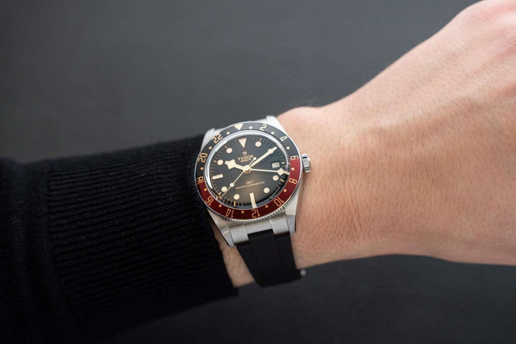 TUDOR Black Bay 58 GMT am schwarzen Kautschukarmband abgebildet an einem Handgelenk mit schwarzem Ärmel vor dunklem Hintergrund