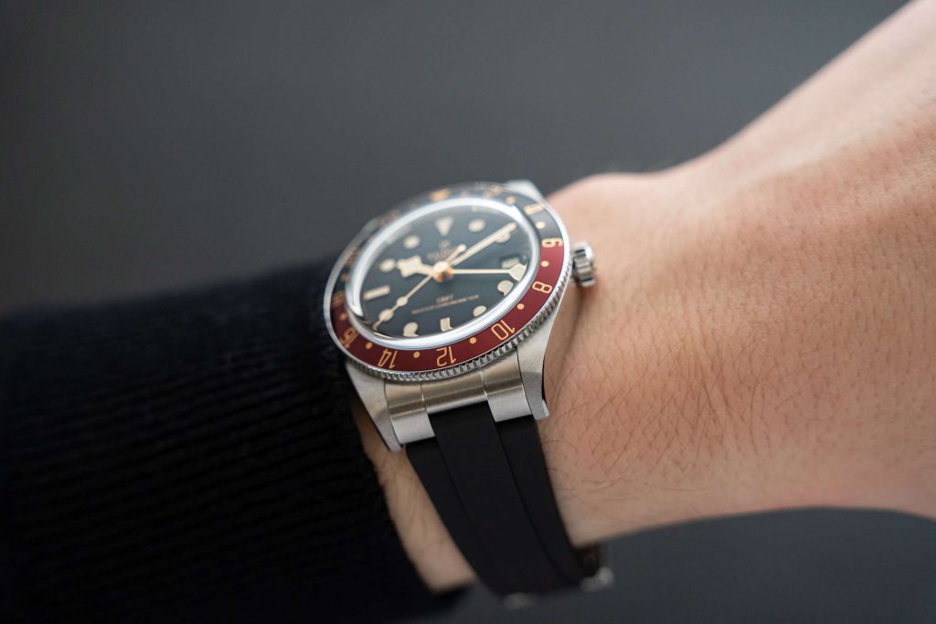 TUDOR Black Bay 58 GMT mit schwarzem Kautschukarmband abgebildet an einem Handgelenk mit schwarzem Ärmel vor grauem Hintergrund