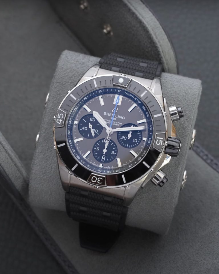Nahaufnahme des Breitling Super Chronomaten B01 Titan am schwarzen Kautschukband um ein Kissen einer Uhrenrolle gebunden vor grauem Hintergrund