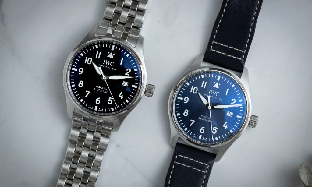 Die IWC Pilot's Watch Mark XX mit schwarzem Zifferblatt am Stahlband links und die IWC Pilot's Watch Mark XX mit blauem Zifferblatt am Lederband rechts liegend vor grauem Hintergrund