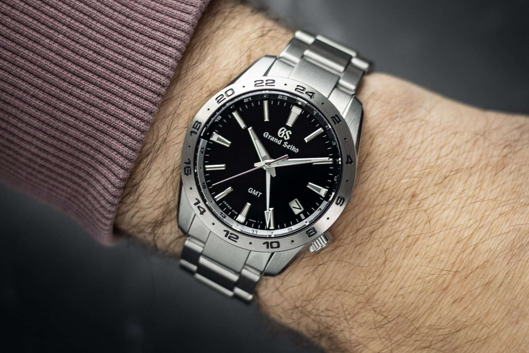 Grand Seiko SBGN027 mit schwarzem Zifferblatt abgebildet an einem Handgelenk mit dunkelrotem Ärmel vor grauem Hintergrund