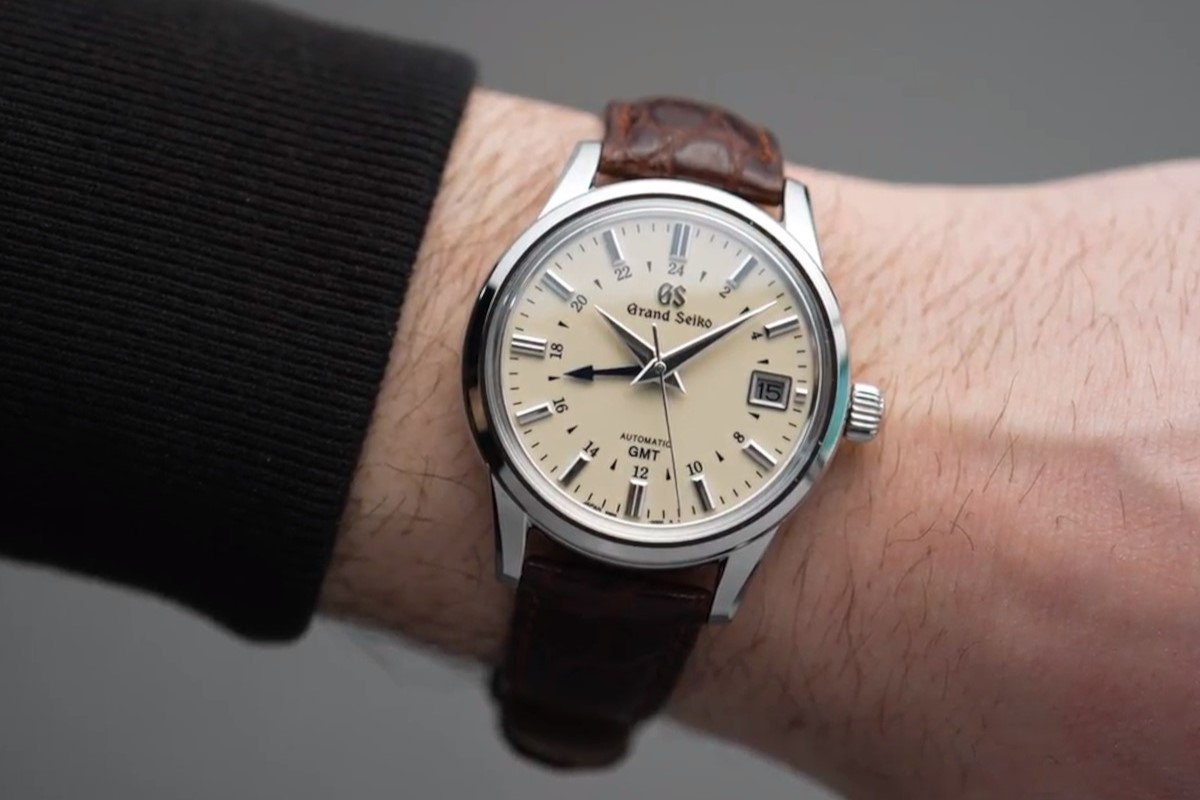 Grand Seiko SBGM221 mit beigem Zifferblatt und braunem Lederarmband abgebildet an einem Handgelenk mit schwarzem Ärmel vor grauem Hintergrund