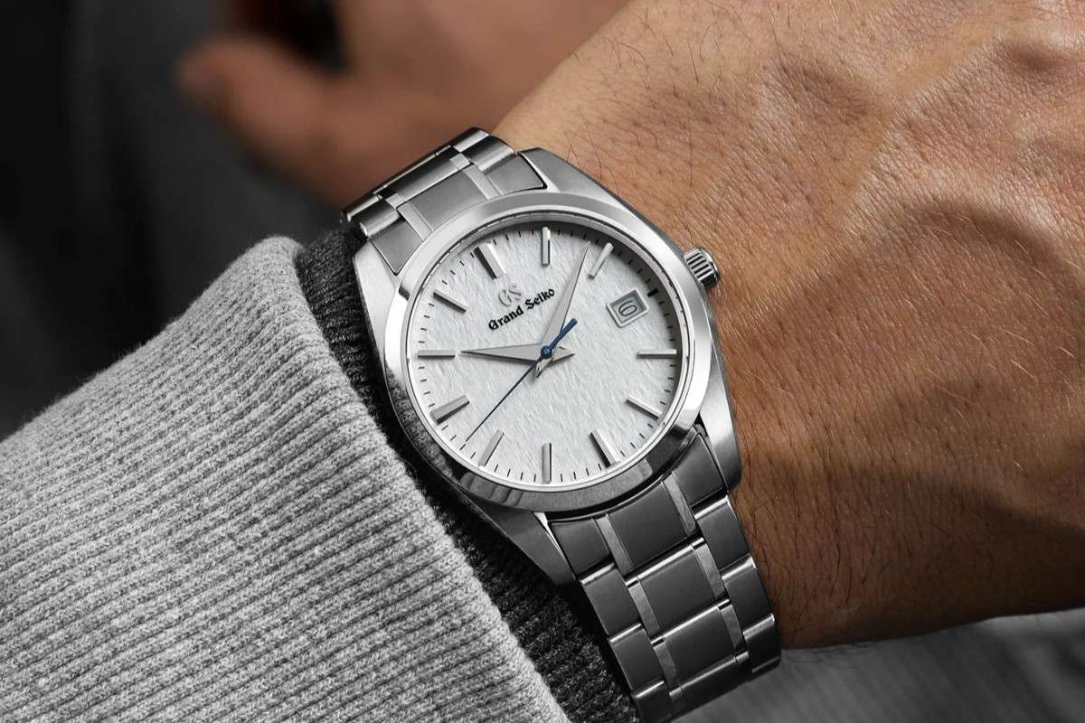 Grand Seiko SBGX355 am Titanarmband mit weißem Zifferblatt abgebildet an einem Handgelenk mit grauem Ärmel vor dunklem Hintergrund