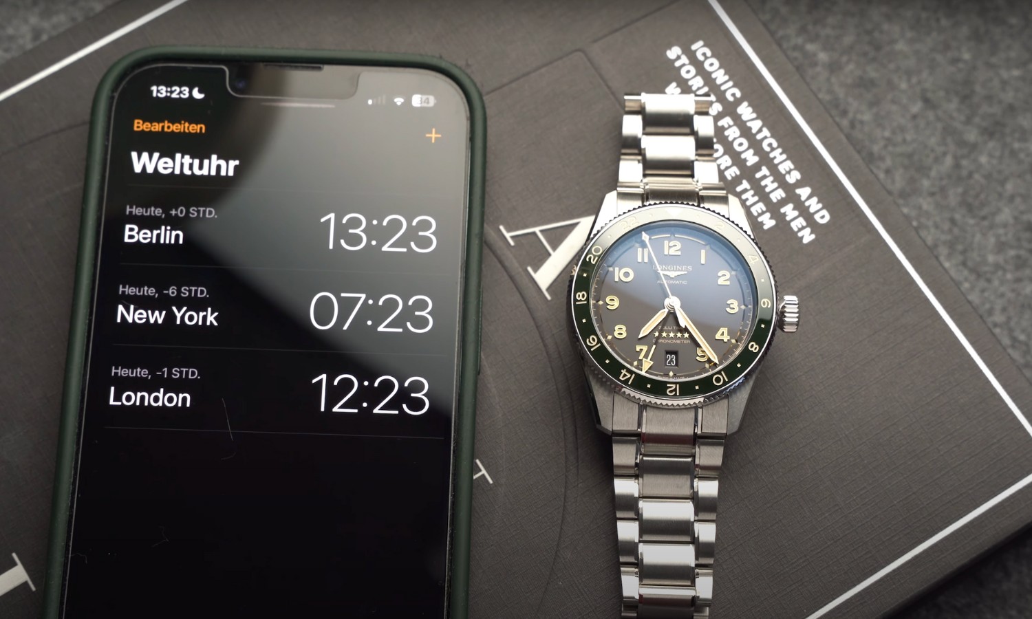 Die Longines Spirit Zulu Time 39 mit schwarzem Zifferblatt am Edelstahlarmband liegend neben einem Handy mit der Anzeige verschiedener Uhrzeiten vor grauem Hintergrund