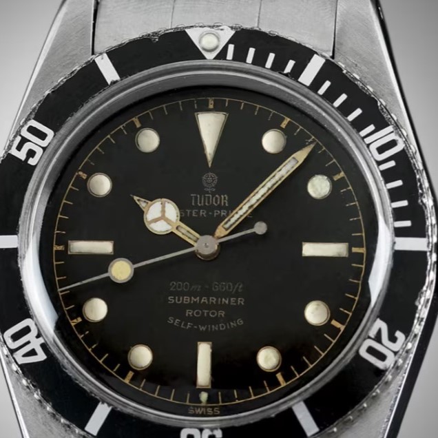 Tudor Oyster Prince Submariner, auch Big Crown genannt aus 1958 mit schwarzem Zifferblatt und schwarzer Lünette am Edelstahlarmband vor grauem Hintergrund