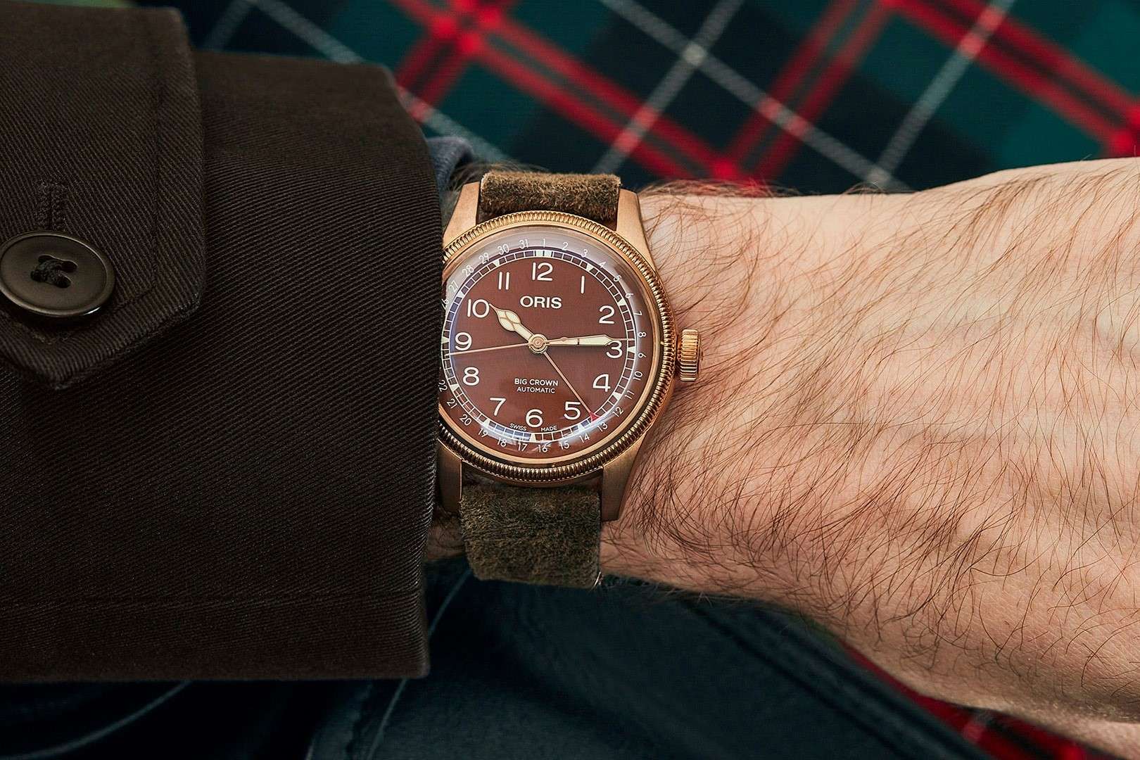 Oris Big Crown Bronze Pointer Date mit braunem Zifferblatt am braunen Lederarmband abgebildet an einem Handgelenk mit dunklem Ärmel vor kartiertem Hintergrund