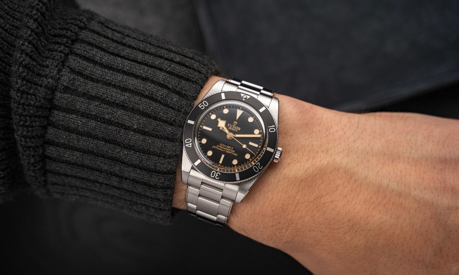 TUDOR Black Bay 54 mit schwarzem Zifferblatt am Edelstahlarmband an einem Handgelenk mit schwarzem Ärmel getragen vor schwarzem Hintergrund