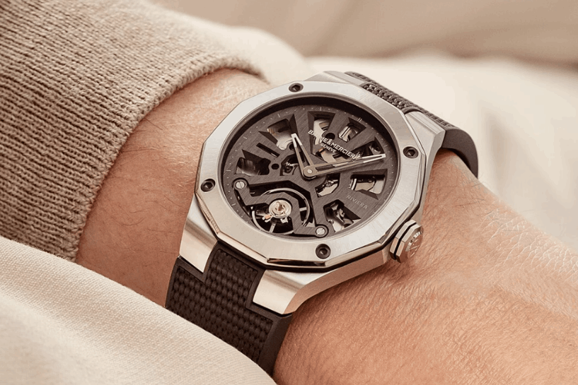 Baume & Mercier Reviera mit skelettiertem Zifferblatt am schwarzen Kautschukarmband abgebildet an einem Handgelenk mit beigem Ärmel vor beigem Hintergrund