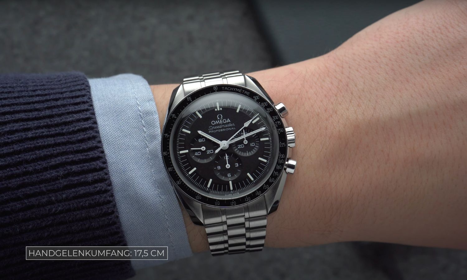 Die OMEGA Speedmaster Professional Moonwatch mit schwarzem Zifferblatt am Edelstahlarmband an einem Handgelenk mit schwarz blauem Ärmel getragen vor grauem Hintergrund