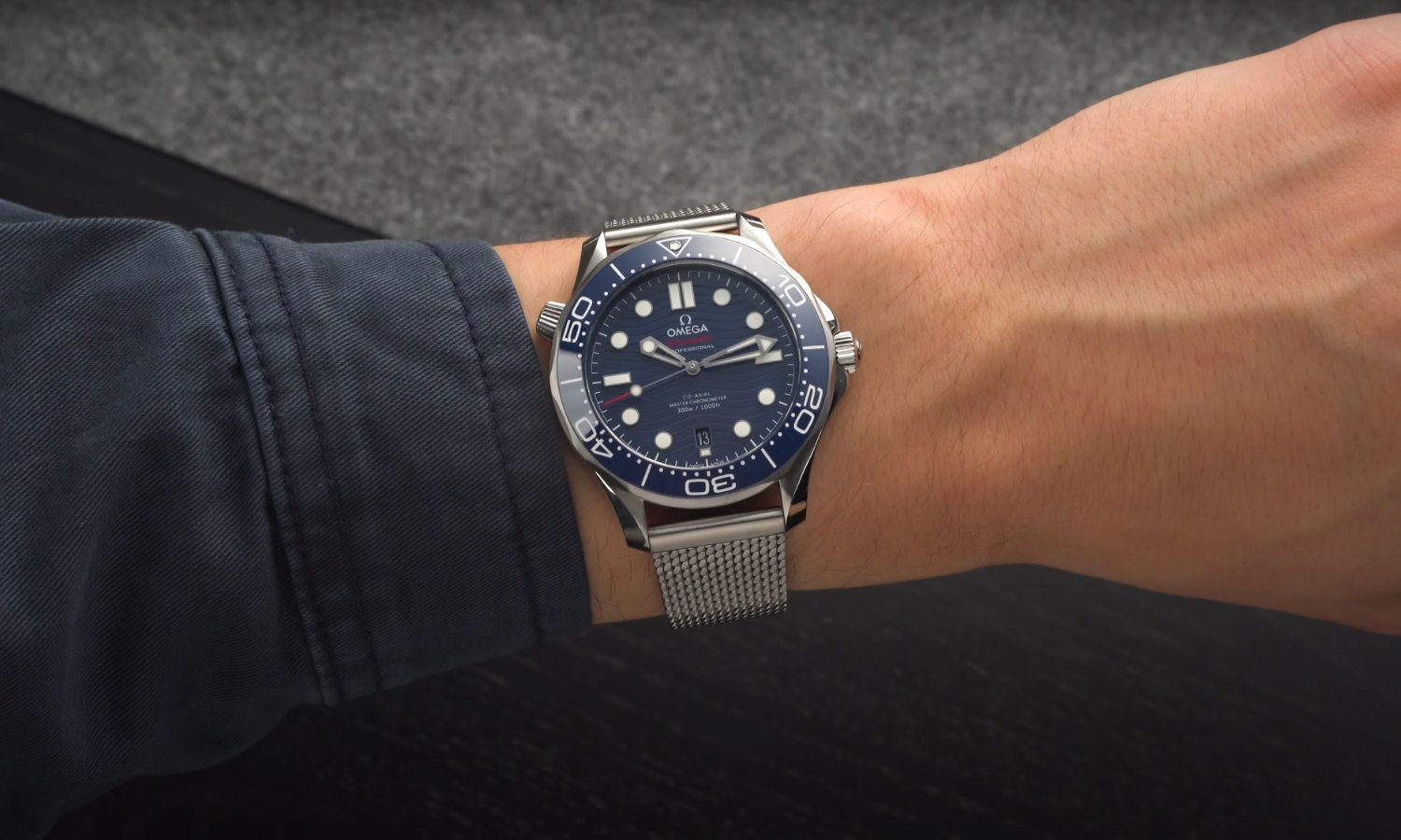 Die OMEGA Seamaster Diver 300M mit blauem Zifferblatt am Edelstahlarmband an einem Handgelenk mit dunkelblauem Ärmel getragen vor schwarz grauem Hintergrund