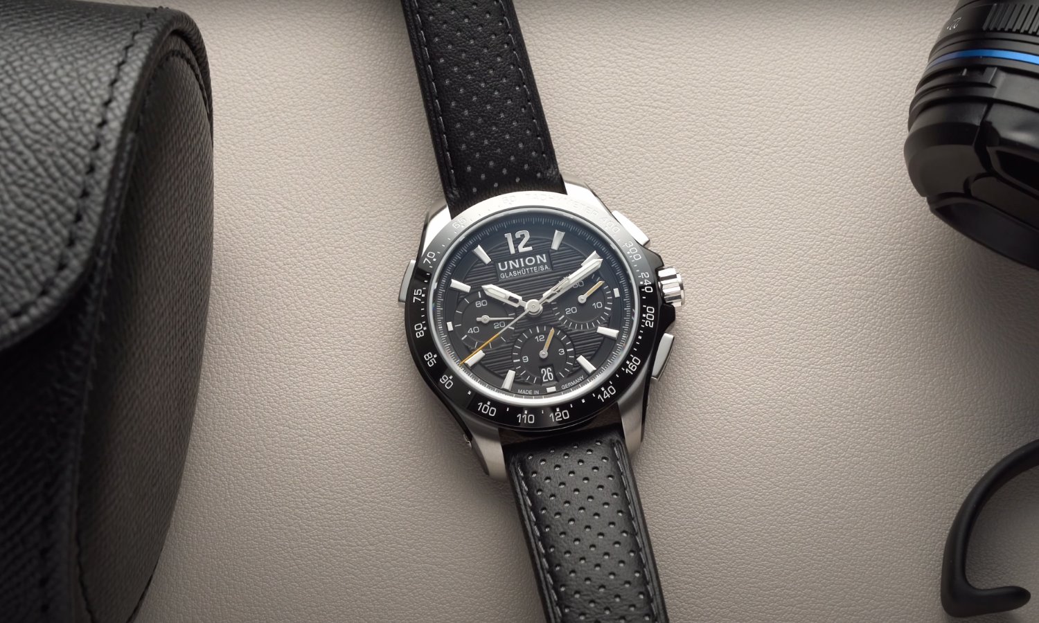 Die UNION Glashütte Belisar Chronograph Sport mit schwarzem Zifferblatt am schwarzen Lederarmband liegend vor hellgrauem Hintergrund neben einer schwarzen Leder-Uhrenrolle