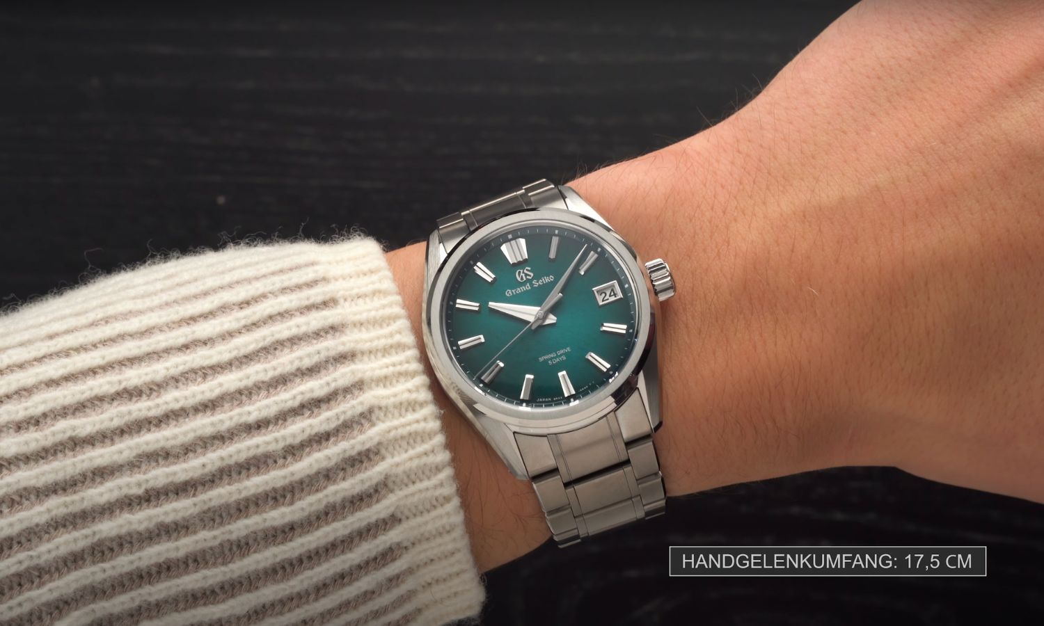 Die Grand Seiko Atera-Valley am Stahlband an einem Handgelenk mit beigem Ärmel getragen vor dunklem Hintergrund