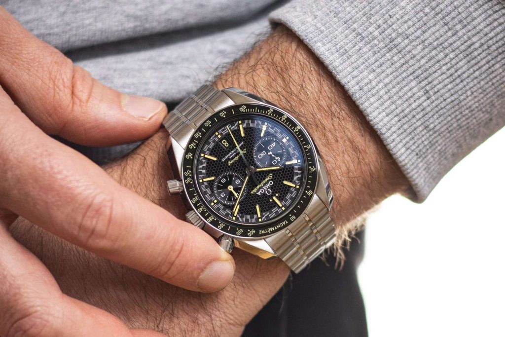 Omega Speedmaster Super Racing abgebildet an einem Handgelenk mit grauem Ärmel vor weißem Hintergrund. Zu sehen sind auch zwei Finger, von denen einer den oberen Chrono-Pusher betätigt