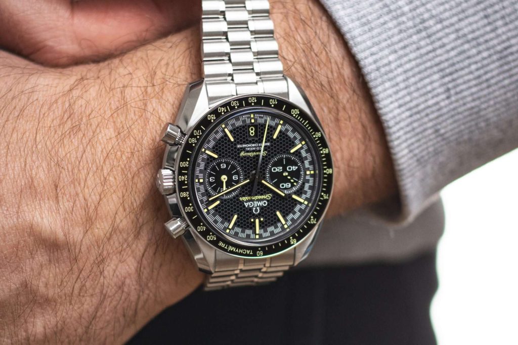 Omega Speedmaster Super Racing abgebildet mit geöffnetem Edelstahlarmband an einem Handgelenk mit grauem Ärmel vor weißem Hintergrund