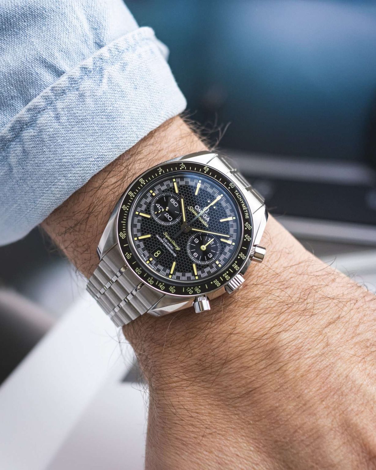 Omega Speedmaster Super Racing am Edelstahlarmband abgebildet an einem Handgelenk mit hellblauem Ärmel vor weißblauem Hintergrund