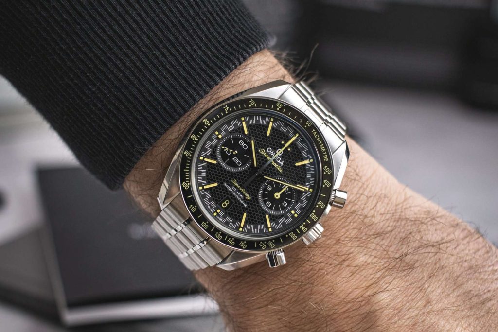 Omega Speedmaster Super Racing abgebildet an einem Handgelenk mit schwarzem Ärmel vor einem hellen Hintergrund mit schwarzem Buch