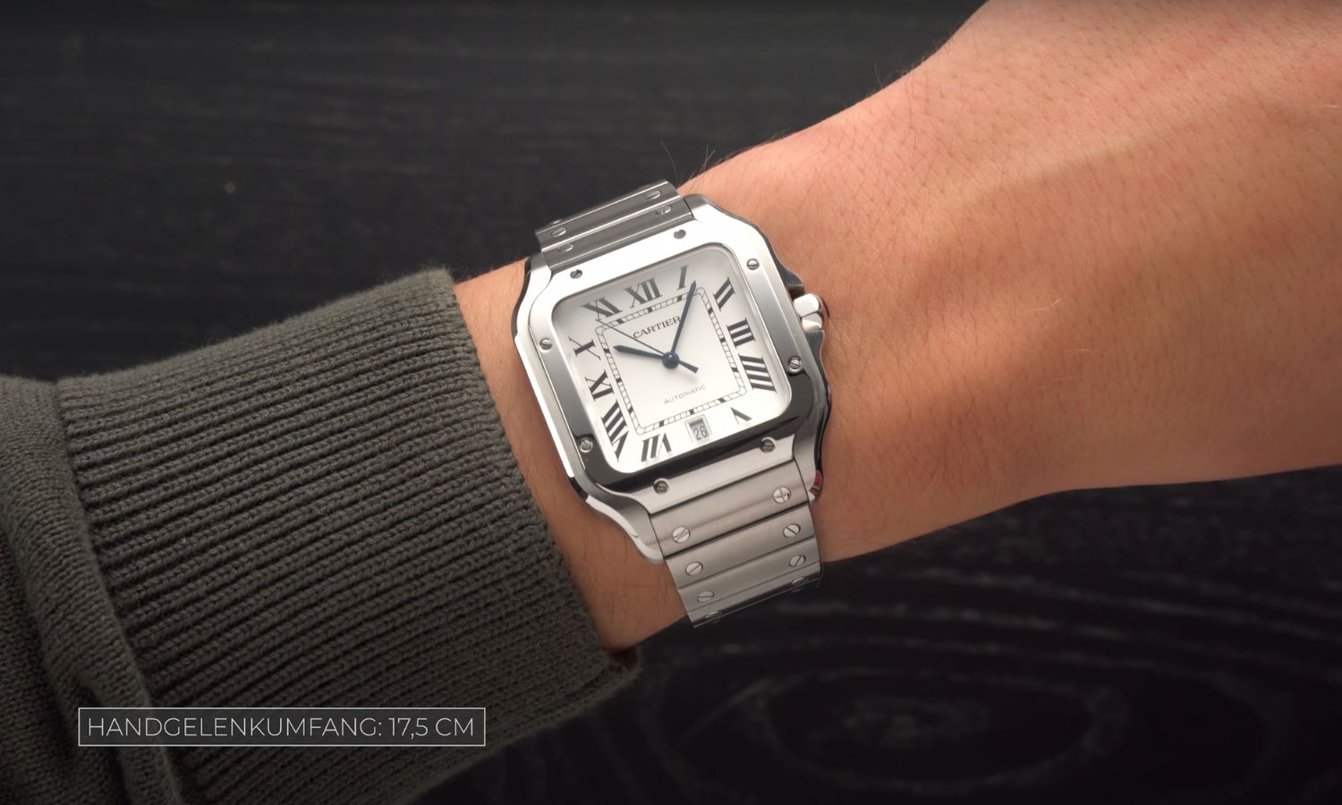 Die Cartier Santos mit weißem Zifferblatt am Edelstahlarmband an einem Handgelenk mit grünem Ärmel vor grauem Hintergrund