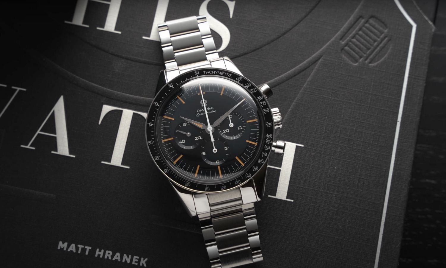 Die OMEGA Speedmaster First OMEGA in Space mit dunkelblauem Zifferblatt am Edelstahlarmband liegend vor schwarzem Hintergrund mit weißem Schriftzug