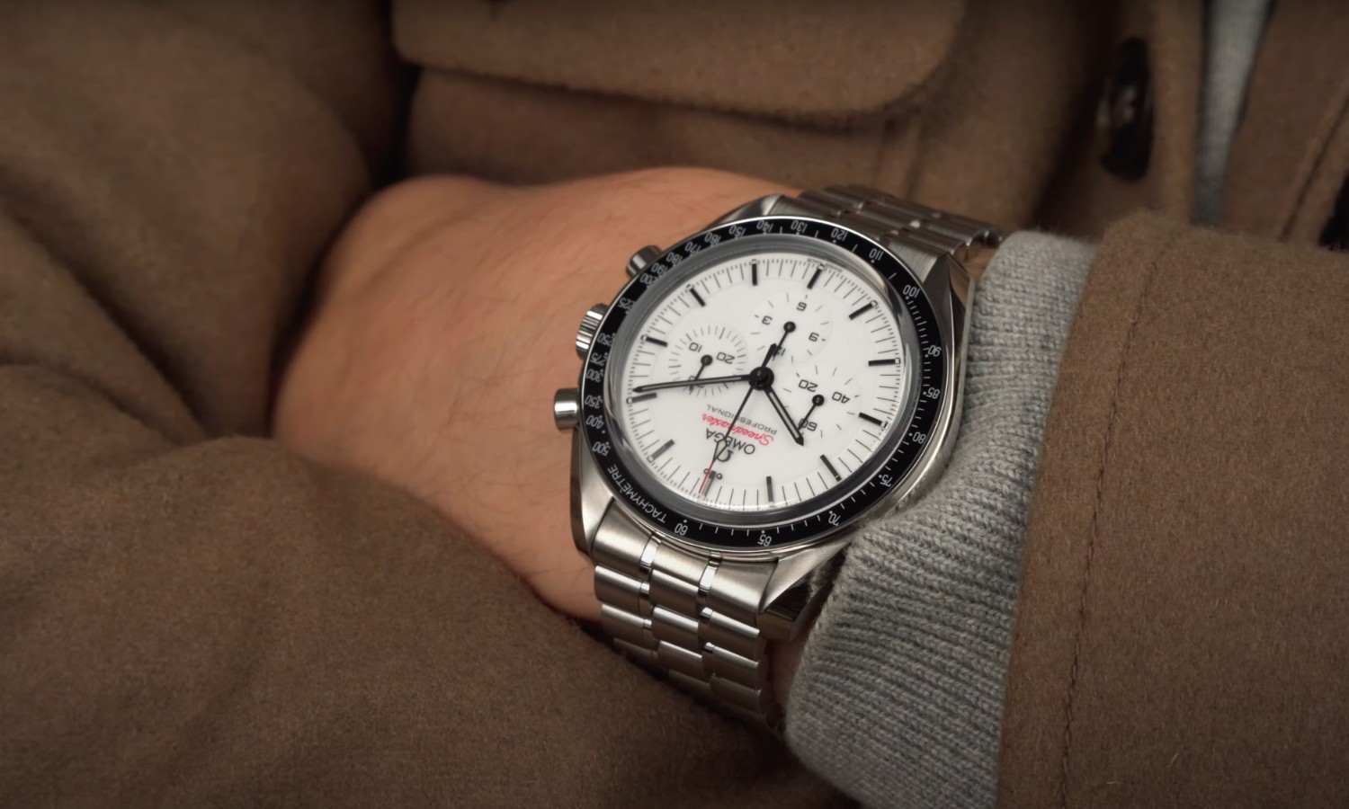 Die OMEGA Speedmaster Professional Moonwatch mit weißem Zifferblatt am Edelstahlarmband an einem Handgelenk getragen mit braunem Ärmel