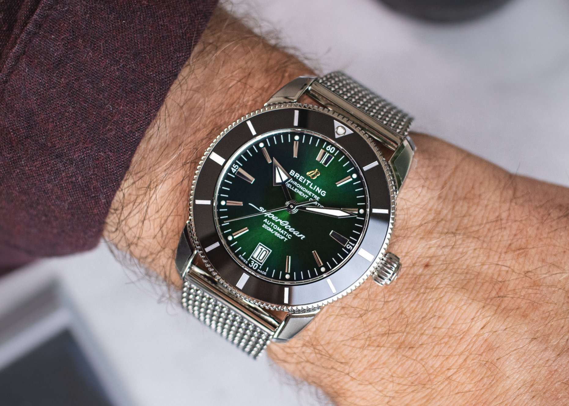 Breitling Superocean Heritage mit grünem Zifferblatt am Edelstahlarmband abgebildet an einem Handgelenk mit dunkelrotem Ärmel vor weißem Hintergrund