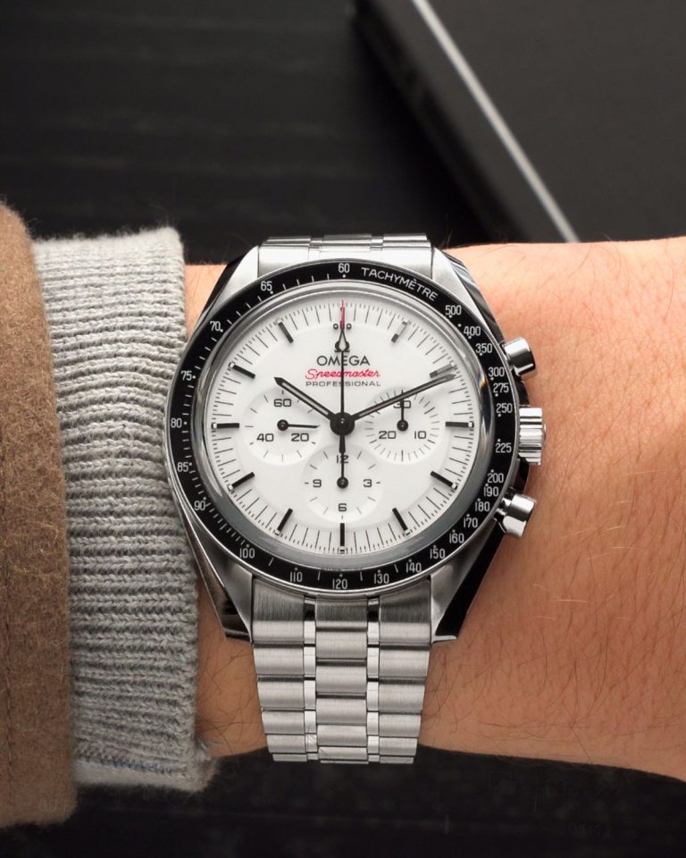 Die OMEGA Speedmaster Professional Moonwatch mit weißem Zifferblatt am Edelstahlarmband an einem Handgelenk mit grau-braunem Ärmel getragen vor schwarzem Hintergrund