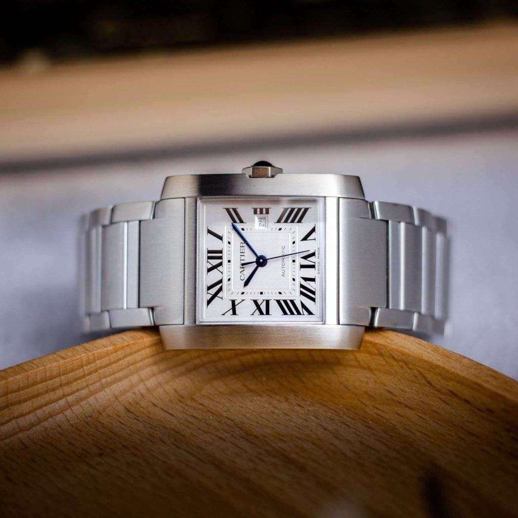 Cartier Tank Francaise mit weißem Zifferblatt und Edelstahlarmband liegend auf einer Holzschale vor weißbraunem Hintergrund