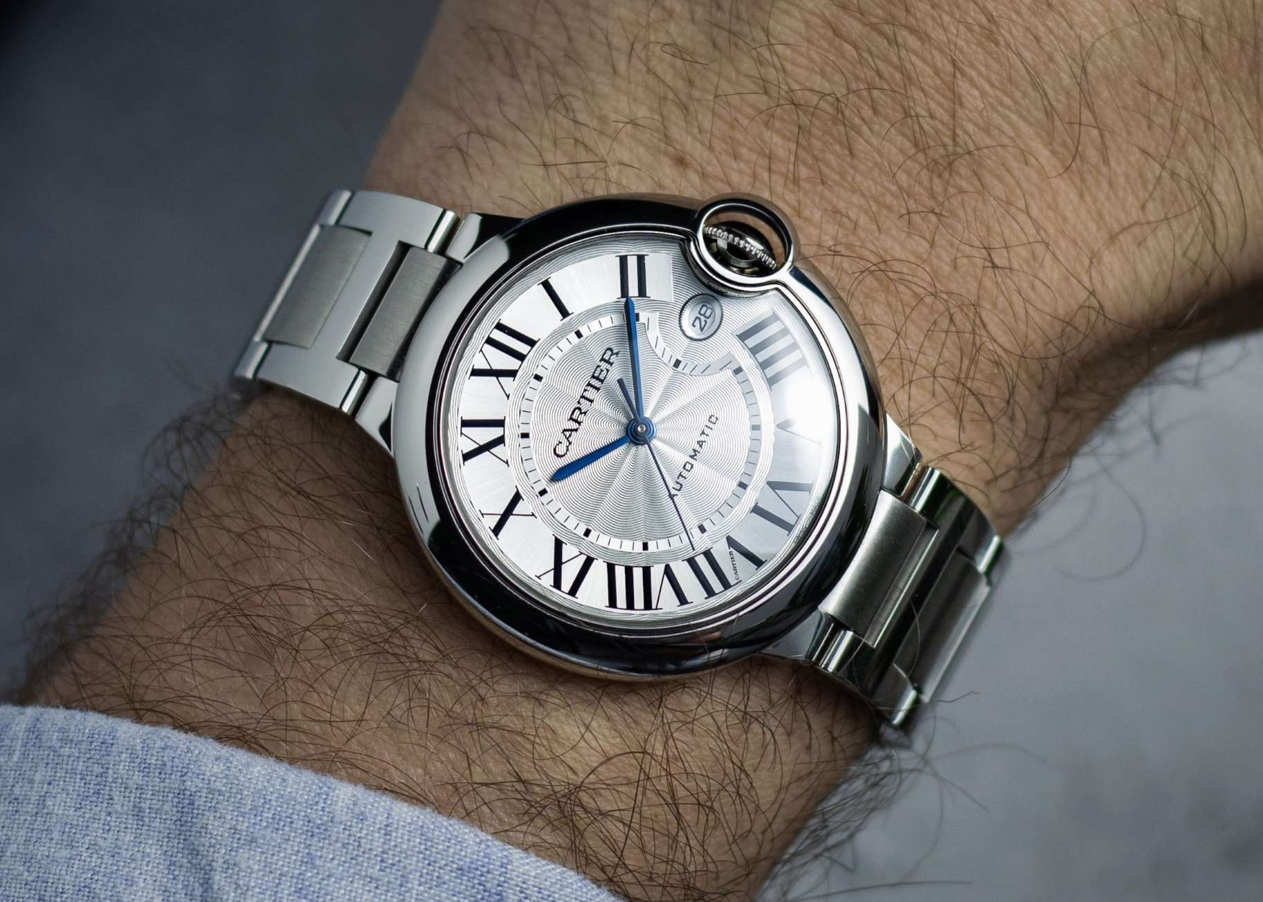 Cartier Ballon Bleu am Edelstahlarmband mit silbernem Zifferblatt abgebildet an einem Handgelenk vor grauem Hintergrund