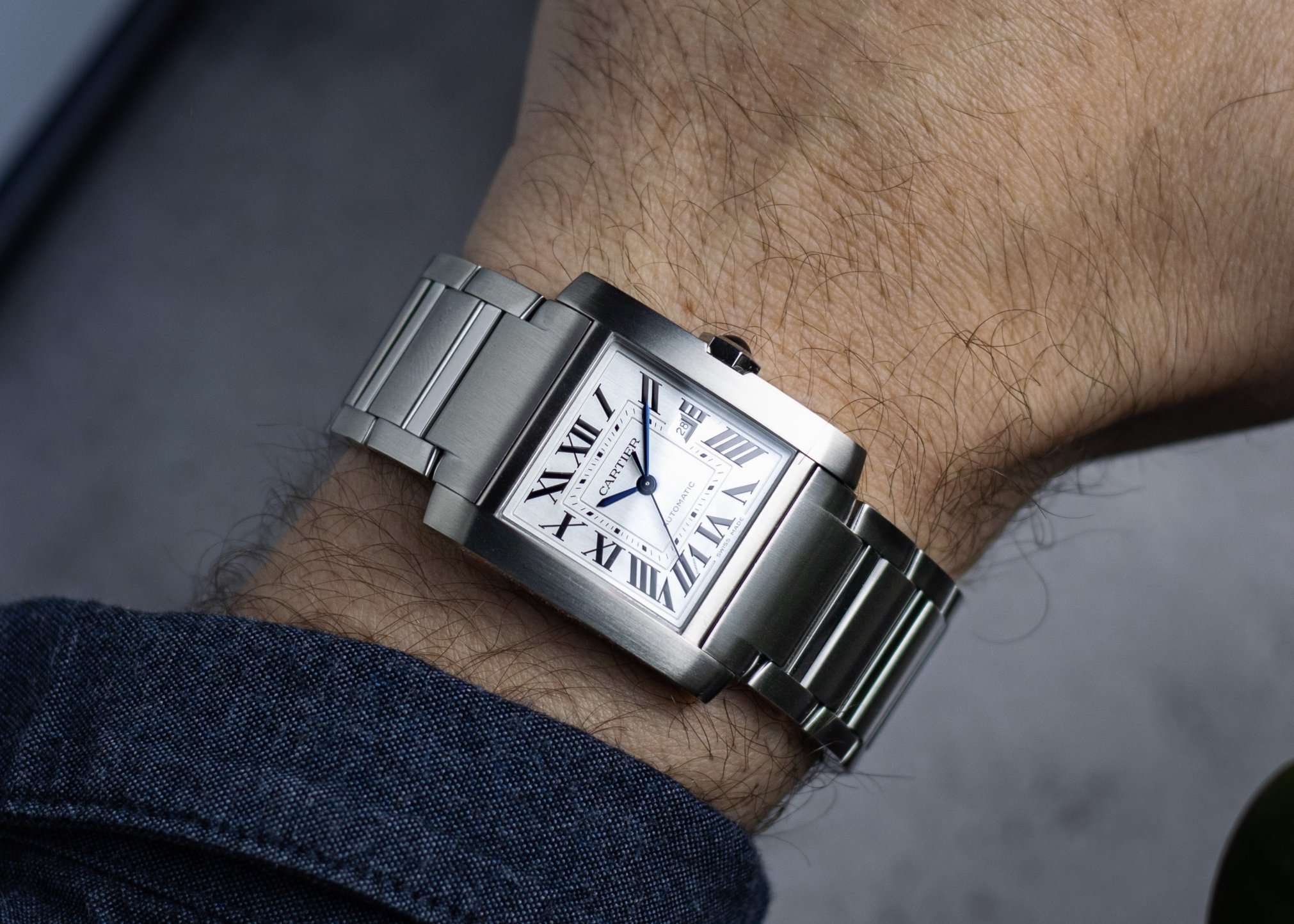 Cartier Tank Francaise mit weißem Zifferblatt und Edelstahlarmband abgebildet an einem Handgelenk mit dunkelblauem Ärmel vor grauem Hintergrund