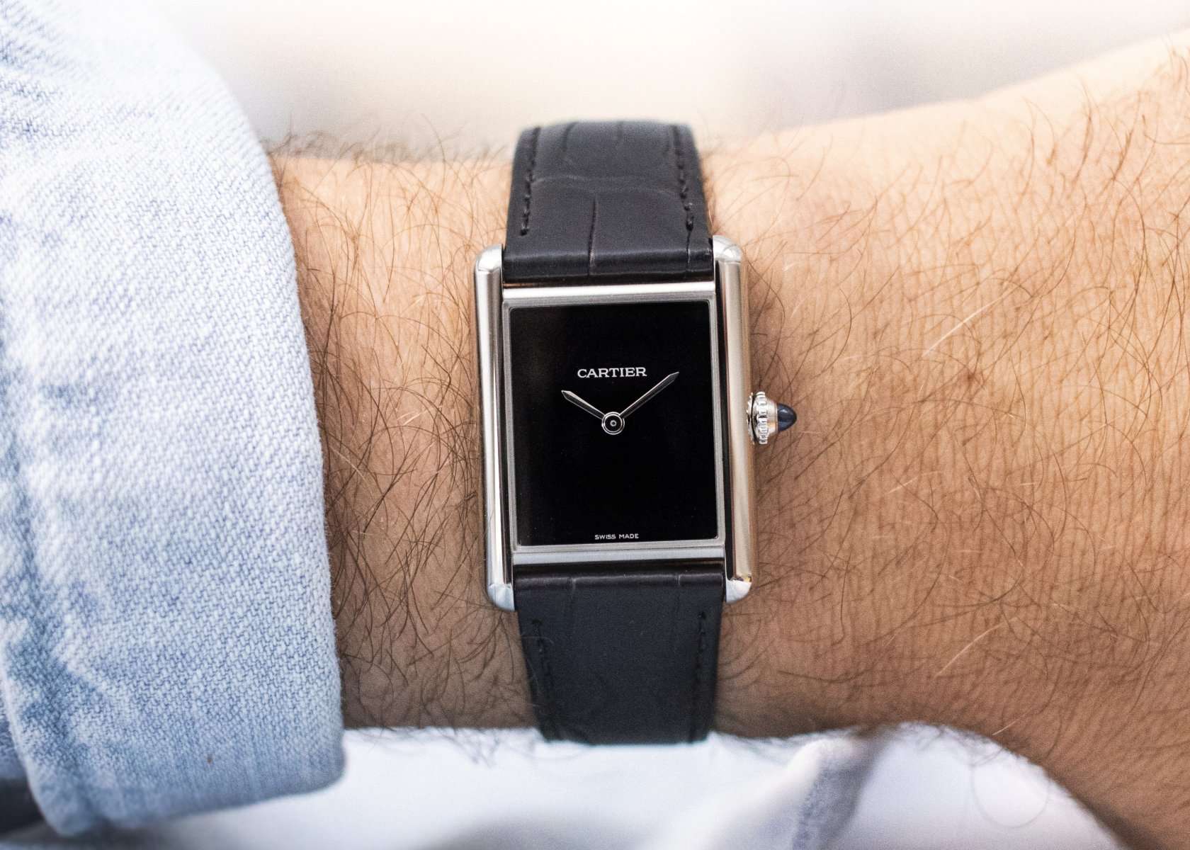 Cartier Tank Must mit schwarzem Zifferblatt und schwarzem Lederarmband abgebildet an einem Handgelenk mit hellblauem Ärmel vor weißem Hintergrund