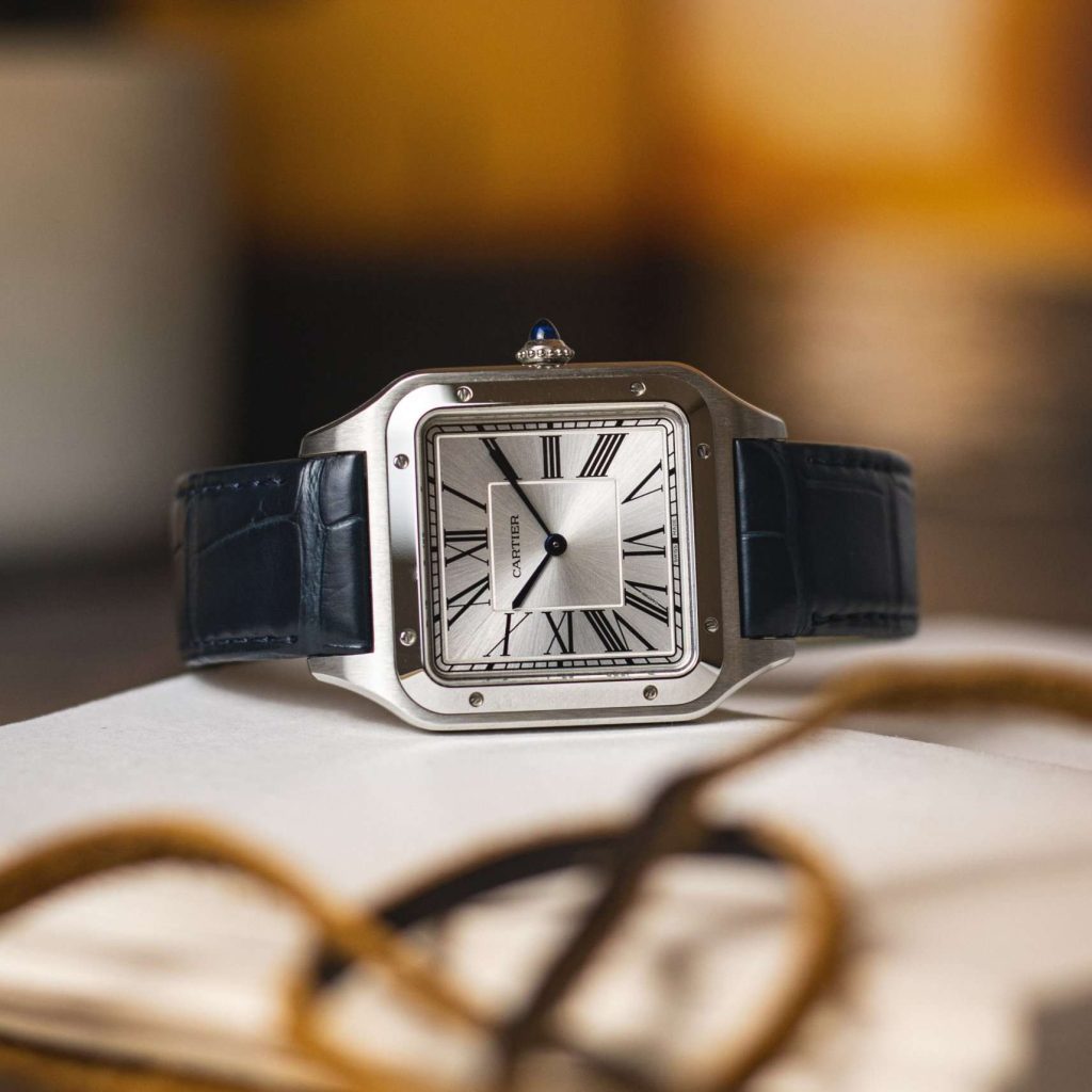 Cartier Santos-Dumont mit silbernem Zifferblatt und blauem Lederarmband liegend auf einem Buch mit Schnüren im Vordergrund und braunem Hintergrund